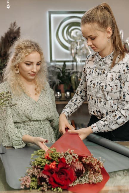 A florist woman with light brown hair tied back, wearing a patterned blouse, carefully wraps a vibrant bouquet of red roses, pink carnations, and white baby's breath in red tissue paper. She is arrang