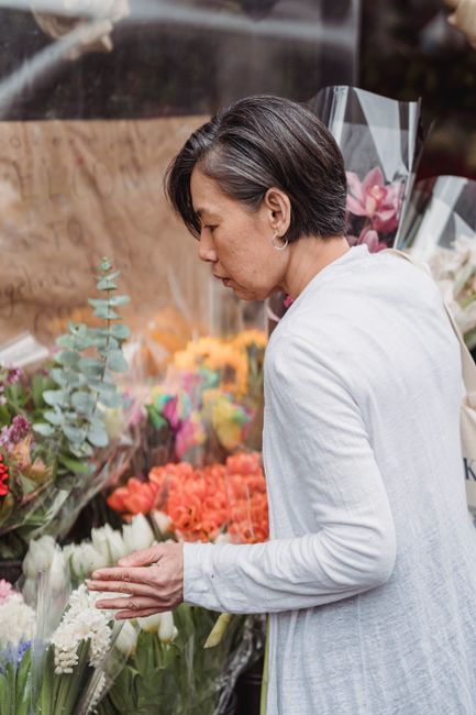 A woman with short dark hair, dressed in a light gray blazer, is observing a display of fresh flowers at a florist shop. The bouquet arrangement in front of her features a variety of flowers, includin