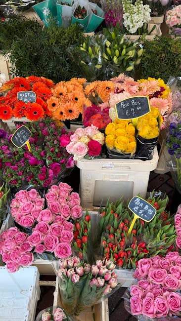 A vibrant display of freshly arranged floral bouquets and potted plants at a flower market, including orange gerbera daisies, peach chrysanthemums, pink roses, yellow roses in a black bucket, and red 