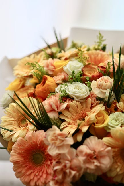 A close-up of a vibrant floral arrangement featuring soft peach and cream gerbera daisies, pale pink lisianthus, white roses, and sprigs of greenery, including long, slender leaves and small fern-like