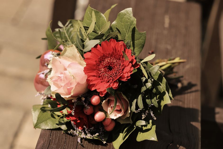 A close-up of a fresh flower bouquet arranged on a wooden surface, featuring a prominent red gerbera daisy at the center, surrounded by soft pink roses, small pink berries, and sprigs of greenery incl
