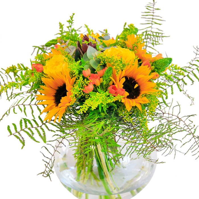 From the heart of Ealing, this radiant bouquet invites you to experience the joyous vibrance of an expertly crafted summer gathering. Sunflowers command attention at the arrangement's center, their bold yellow-orange petals and inky cores reaching outward like tiny suns scattered across the local Gunnersbury landscape. Tucked beside them, plump yellow roses unfold in elegant spirals, bringing a timeless beauty reminiscent of garden strolls along Mattock Lane. Rich red Hypericum berries are artfully nestled among a tapestry of green, offering glimmers of colour that dance playfully past the soft curves of fern and wispy goldenrod. Eucalyptus leaves of subtle blue-green rest between blooms, conjuring both visual contrast and a refreshing herbal note carried on the breeze through Lammas Park. The bouquet overflows from a clear, spherical glass vase whose transparency highlights every twist of stem and shimmer of water, anchoring the composition in simple purity. As natural light pours across a nearby table, the arrangement glows with a sense of celebration, making it perfect for birthday surprises, anniversary tributes, or as a welcoming gesture for close friends gathering in the heart of Ealing. Each flower and leaf speaks of freshness, artistry, and attention to detail, inviting joyful connection with every glance and breath.